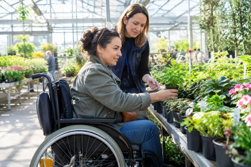 Une femme en fauteuil choisit une plante dans une serre municipale avec une agente.
